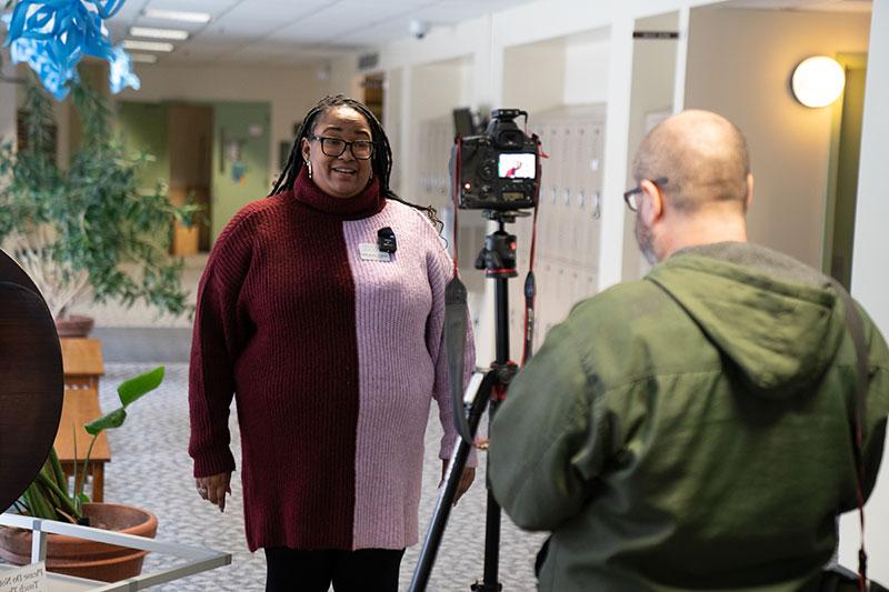 Alicia Lloyd speaking in front of a camera in a hallway while someone operates the camera