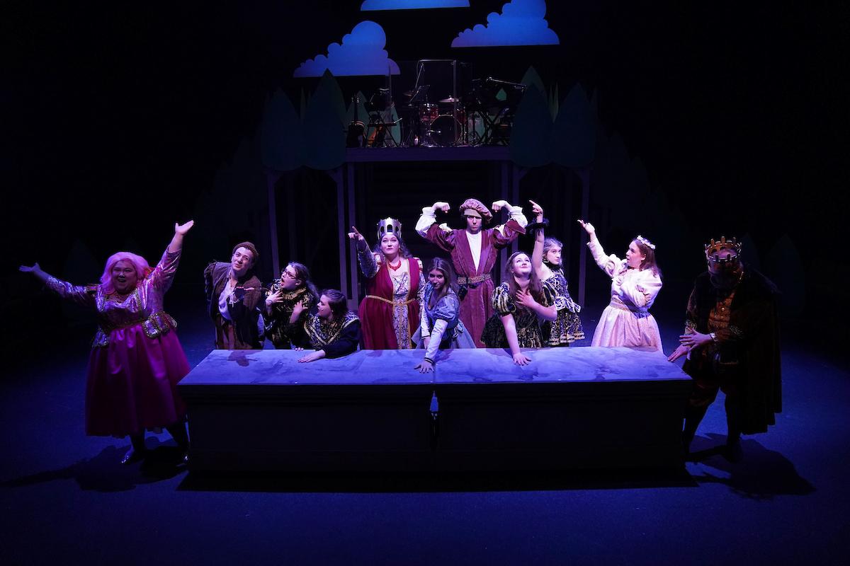 Several actors in various fairytale costumes line one side of the table, striking poses in blue light on the stage 