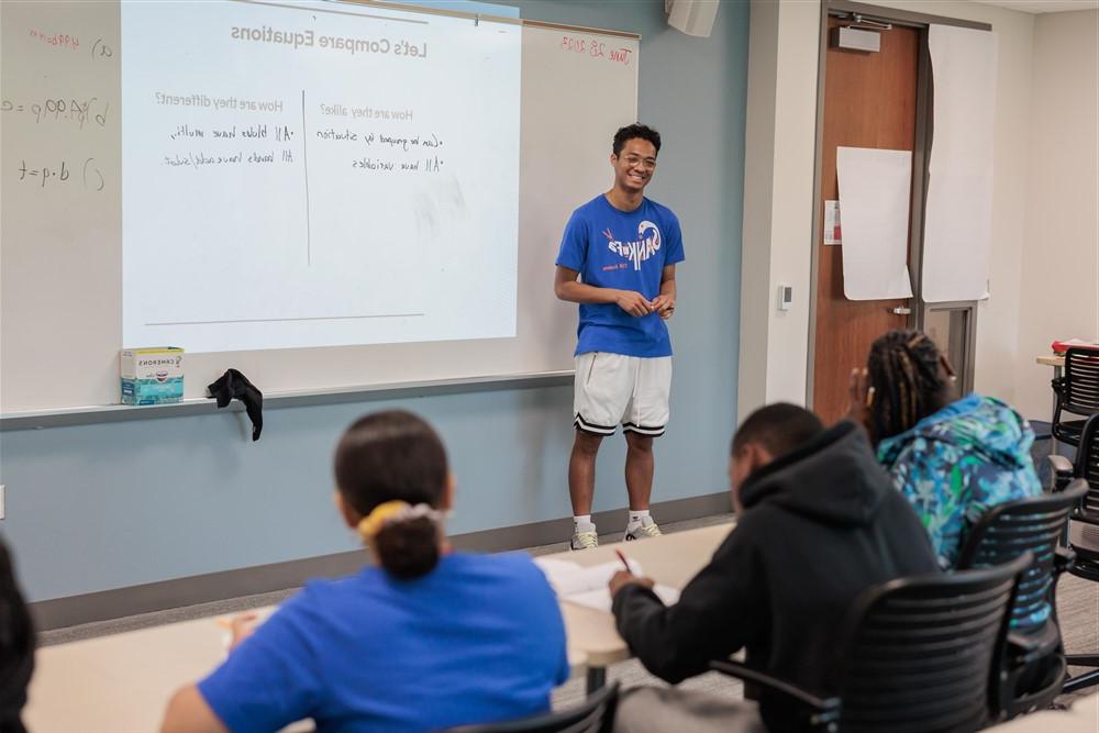 Students listening to a teacher who is smiling in front of a whiteboard